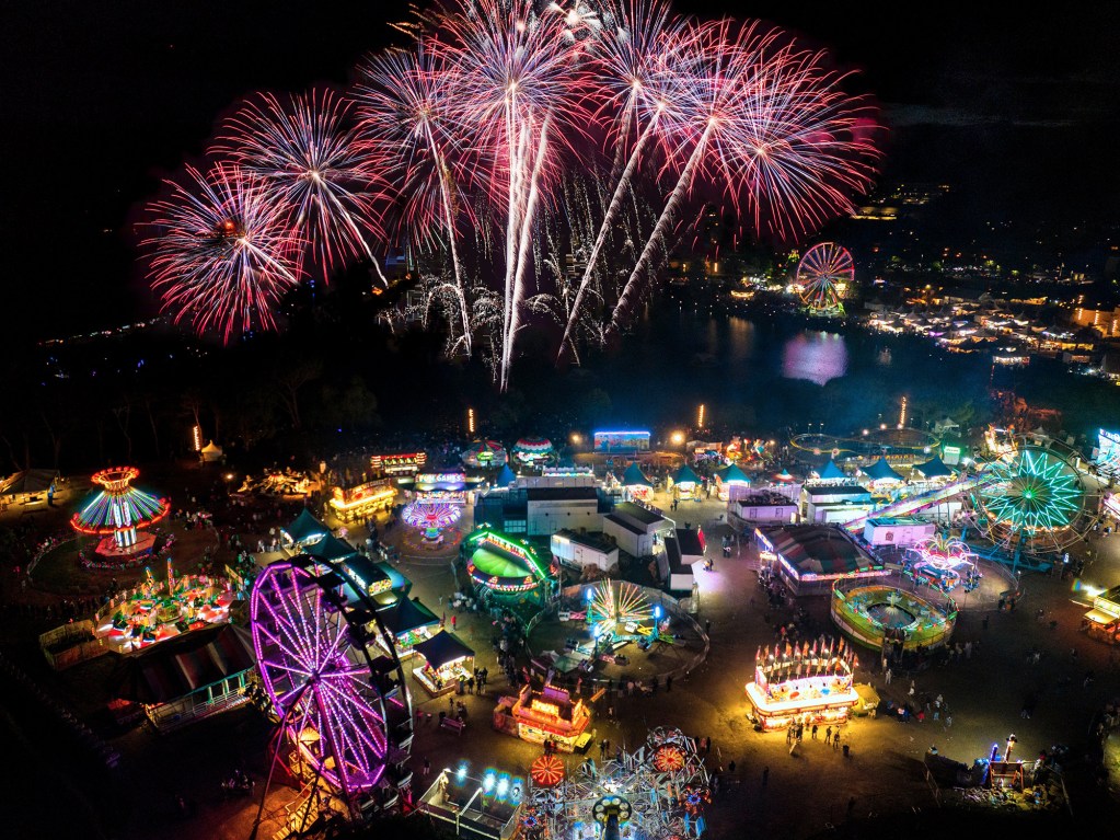 Aerial view of a vibrant carnival at night, featuring colorful rides, games, and food stalls, illuminated by fireworks in the sky.