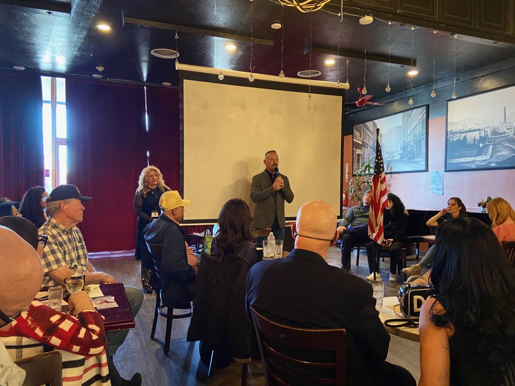A California gubernatorial candidate, Sheriff Chad Bianco, addresses an audience in a coffee shop. Attendees listen attentively, with an American flag visible in the background.
