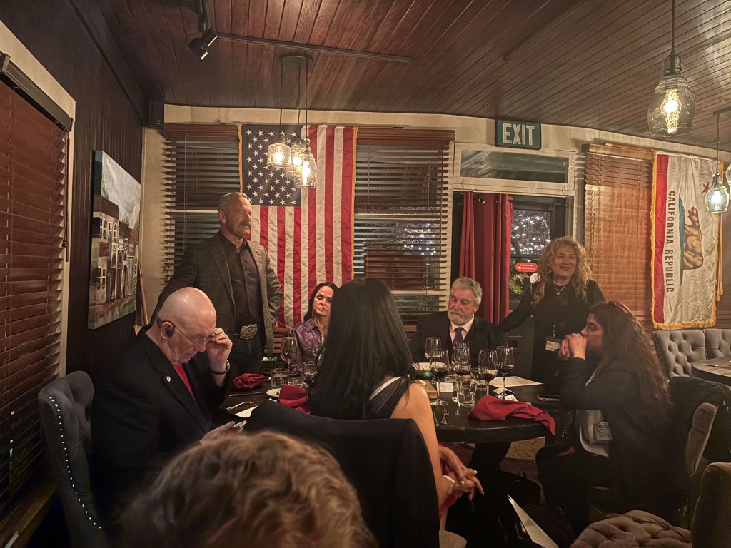 A group of people seated at a dinner table, with one man standing and speaking, an American flag hanging in the background, and a California state flag visible.