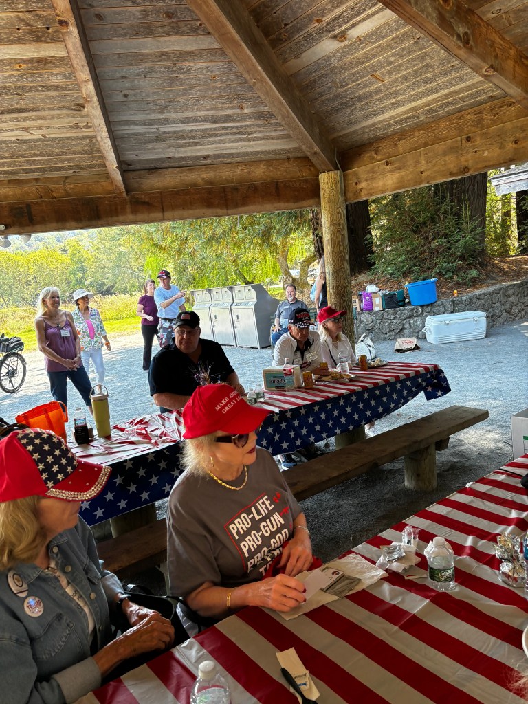 Group of people gathered under a wooden pavilion with American flag-themed table covers, engaging in a discussion or event, with some wearing red hats and themed attire.