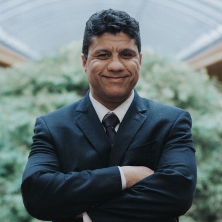 A smiling man in a dark suit with arms crossed, posing against a blurred green background.