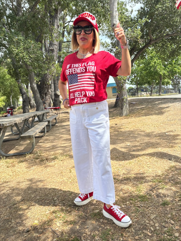 Woman standing outdoors wearing a red shirt with an American flag and text, white pants, and red sneakers, smiling and posing.