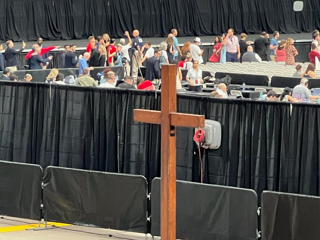 A wooden cross in the foreground with a crowd of people gathered behind black curtains and seats at an event.