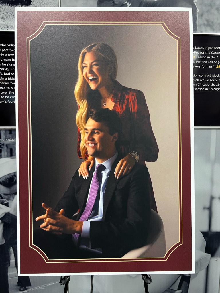 A smiling woman in a red dress is playfully sitting behind a man in a suit, who is seated and smiling. The background is softly lit, highlighting their cheerful expressions.