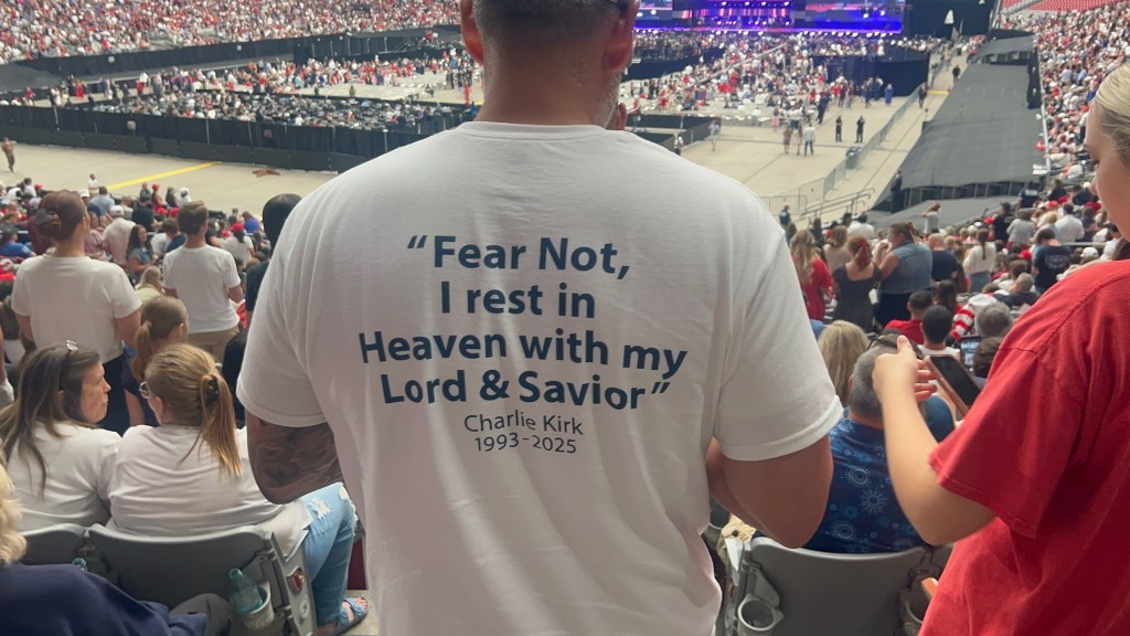A person wearing a white shirt with the text 'Fear Not, I rest in Heaven with my Lord & Savior' and the name Charlie Kirk 1993-2025, standing in a crowded arena.