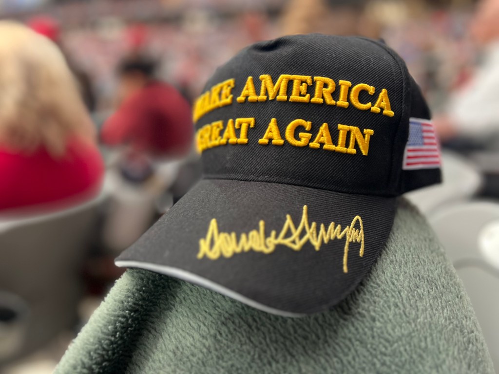 A black 'Make America Great Again' hat with yellow lettering and a signature on the brim, resting on a green fabric in a crowded setting.