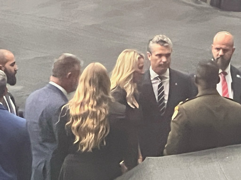 A group of people in formal attire, including a woman with long blonde hair and a man in a suit, engaged in conversation with a uniformed officer in a public setting.