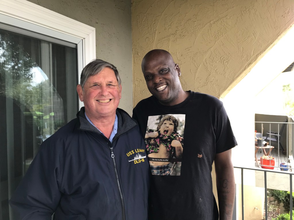 Two men posing together outside, with one wearing a jacket featuring 'USS Leahy' and the other in a black t-shirt with a colorful graphic.